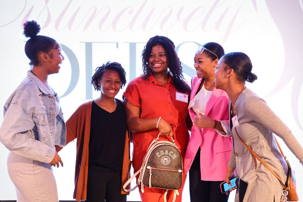 Five young ladies on stage laughing with joy, one is holding a backpack.
